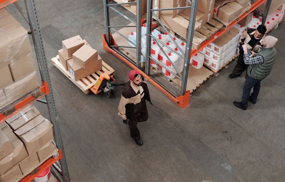 Petugas menarik pallet jack berisi kardus di gudang pelanggan, dua orang lain mencatat stok.