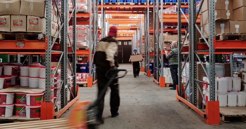 Lorong gudang dengan rak tinggi penuh stok dan petugas memindahkan barang.